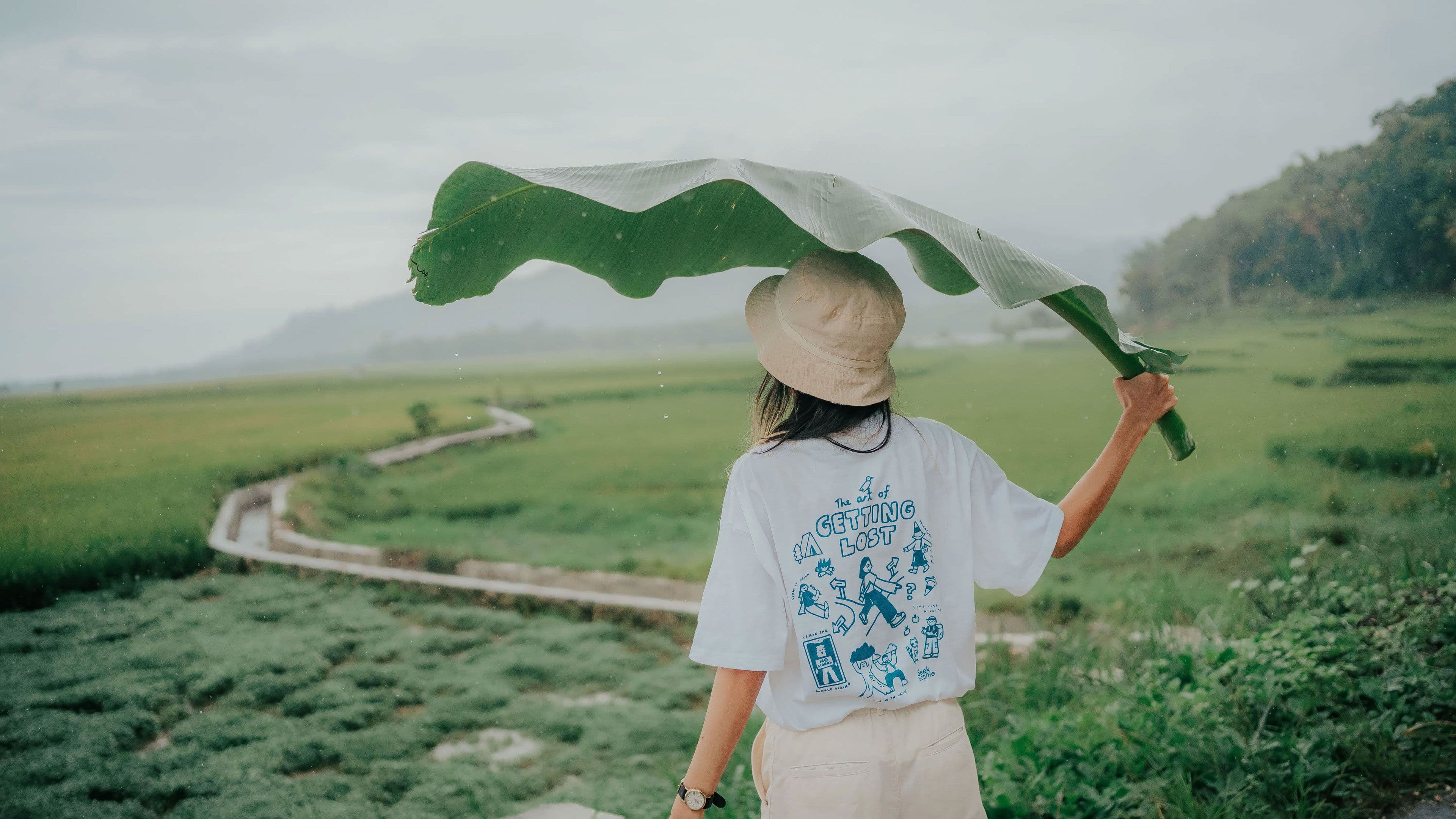 An explorer in a bucket hat, wearing a Seek Sophie "Out of Office" t-shirt and waving a giant leaf above her head. She is staring into the distance, looking out toward a natural landscape in an unknown location